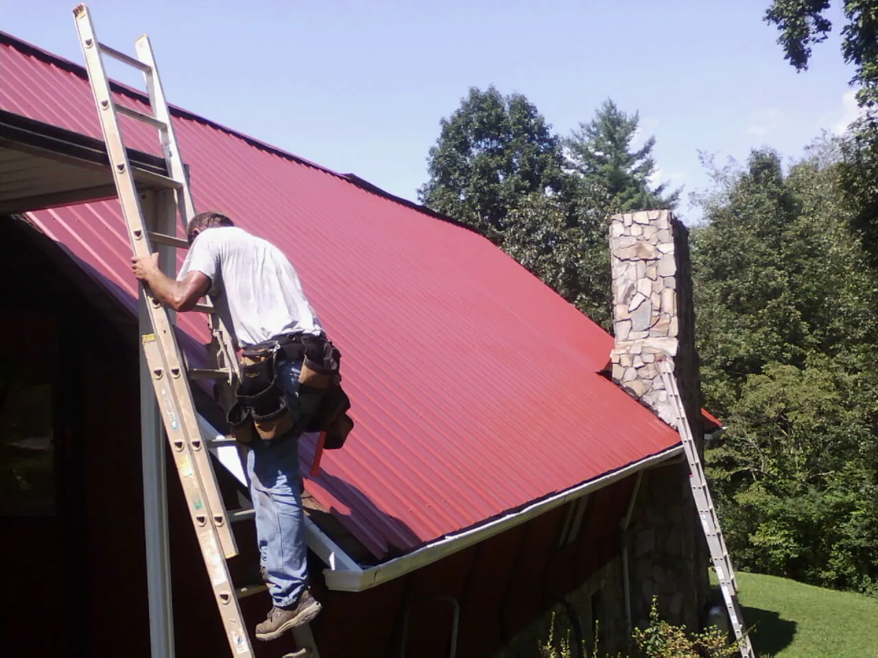 Roofer on a ladder beside a red metal roof and stone chimney — Soffit & Fascia Repair by Roofing Brothers in Sumerduck